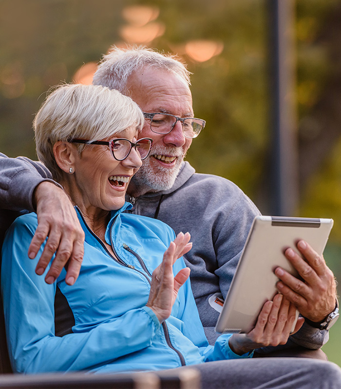 Two seniors outside smiling down at a tablet together on a park bench.